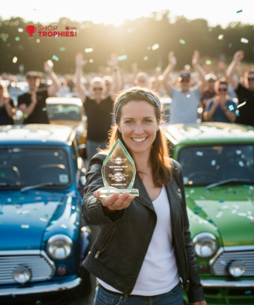 Woman holding an award in front of a crowd and vintage cars with 'Shop Trophies!' branding.