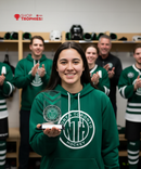 Person in a green hoodie holding a trophy in a hockey locker room with teammates clapping.