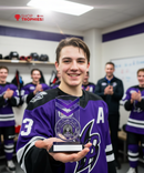 Person in a hockey jersey holding an award in a locker room with teammates and coaches.